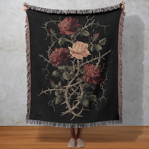 May include: Black throw blanket with a floral design of roses and thorny vines in red, cream, and green. The blanket has a fringed edge and is held up by a person.