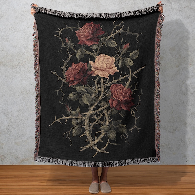 May include: Black throw blanket with a floral design of roses and thorny vines in red, cream, and green. The blanket has a fringed edge and is held up by a person.