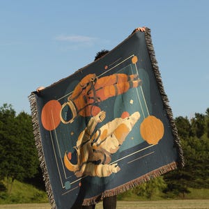 May include: A dark blue throw blanket featuring a space-themed design with two astronauts in orange and beige suits. The blanket has a fringed edge and geometric shapes in orange and white. The background is a blue sky.