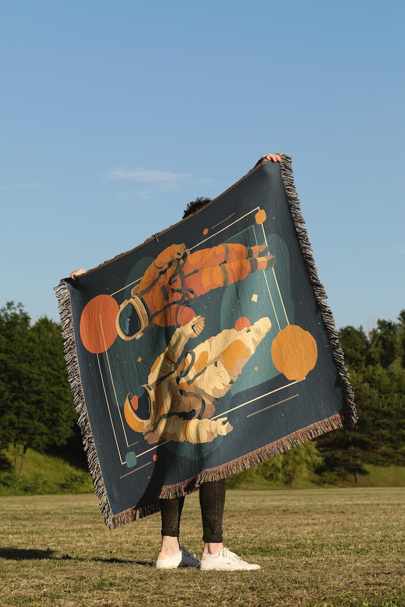 May include: A dark blue throw blanket featuring a space-themed design with two astronauts in orange and beige suits. The blanket has a fringed edge and geometric shapes in orange and white. The background is a blue sky.