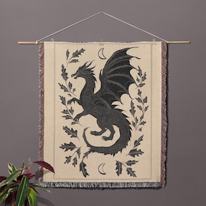 May include: A woven wall tapestry featuring a black dragon design. The dragon is surrounded by leaves and crescent moons, set against a beige background. The tapestry is rectangular with a fringed edge and is hung from a wooden rod.
