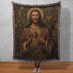 May include: A woven tapestry blanket featuring a religious image of a man with long hair and a halo, holding a flaming heart. The background is a lush, green and brown floral design. The blanket has a fringed edge.