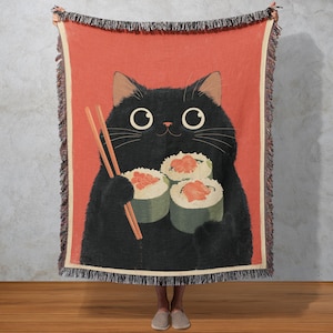 Op de afbeelding: Een sprei met een illustratie van een zwarte kat die sushi en eetstokjes vasthoudt. De achtergrond is rood met een crèmekleurige rand. De kat heeft grote ogen en een lachend gezicht. De sprei heeft een gefranjerde rand.
