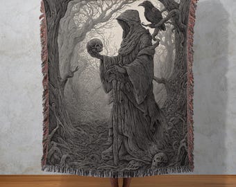 May include: A woven throw blanket with a detailed illustration of a robed figure holding a skull in a dark forest. A raven perches on a tree branch. The artwork is in shades of grey and black, with fringed edges.