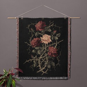 May include: A black tapestry with a botanical illustration of roses and thorns. The roses are in shades of red and cream, with green leaves and thorny vines. The tapestry is hung from a wooden rod and has a fringed edge.