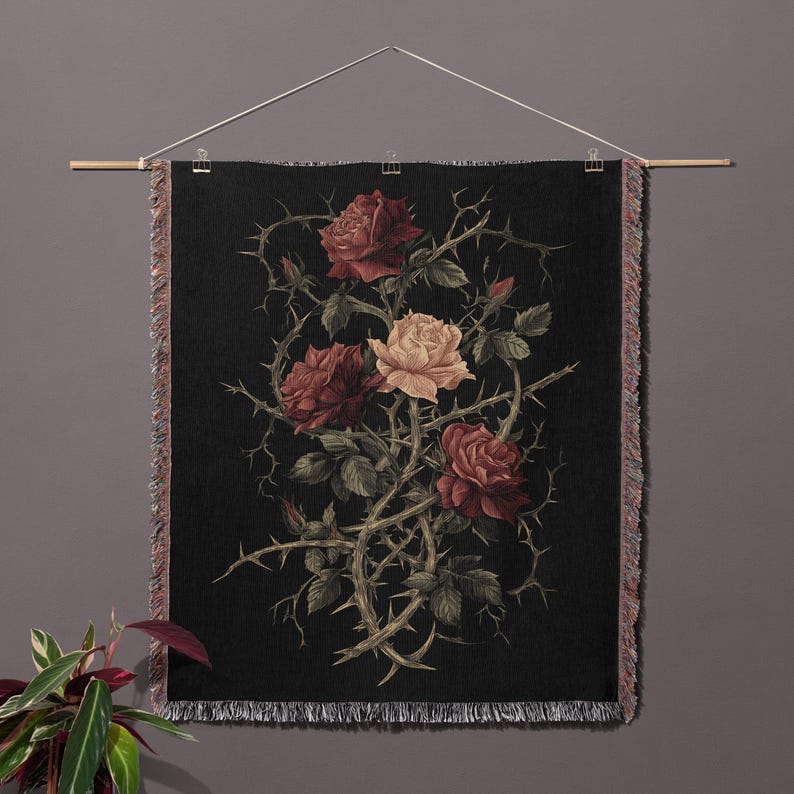 May include: A black tapestry with a botanical illustration of roses and thorns. The roses are in shades of red and cream, with green leaves and thorny vines. The tapestry is hung from a wooden rod and has a fringed edge.