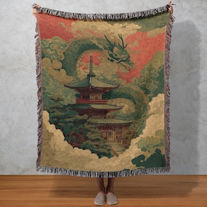 Könnte beinhalten: Gewebte Decke mit japanisch inspiriertem Design. Das Kunstwerk zeigt einen grünen Drachen, eine Pagode und Wolken in Grün-, Rot- und Beigetönen. Die Decke hat Fransen und wird von einer Person gehalten.