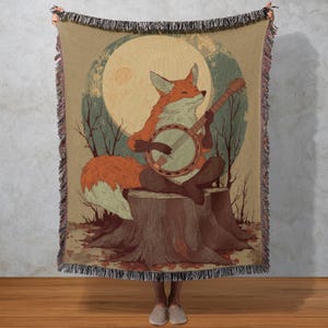 May include: A woven throw blanket featuring a whimsical illustration of a fox playing a banjo. The fox, with orange and white fur, sits on a tree stump against a backdrop of a full moon and bare trees. The blanket has a fringed edge.