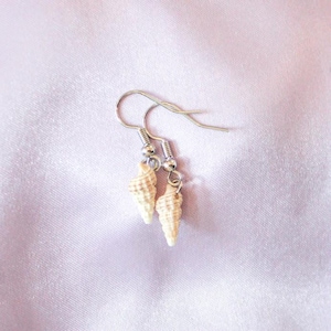 May include: A pair of seashell earrings with silver-colored hooks. The earrings feature small, cream-colored seashells with a spiral design. The background is a light pink, silky fabric.