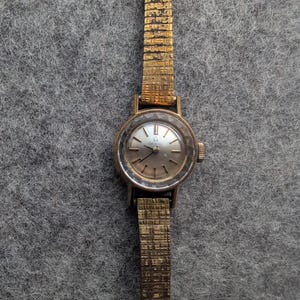 May include: A vintage gold-toned wristwatch with a textured band and a round face. The watch face has a silver-colored background with gold-toned hands and hour markers. The watch band is approximately 0.5 inches wide.