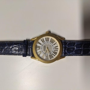 May include: A gold-tone Guess watch with a white face and Roman numeral hour markers. The watch has a blue leather strap with a crocodile pattern.