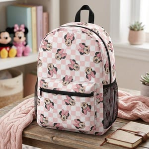 Peut inclure: Un sac &agrave; dos avec un motif r&eacute;p&eacute;titif de visages de Minnie Mouse avec des n&oelig;uds roses sur un fond &agrave; carreaux rose clair et blanc. Le sac comprend une poign&eacute;e sup&eacute;rieure, une poche avant zipp&eacute;e et des poches lat&eacute;rales en filet noir.