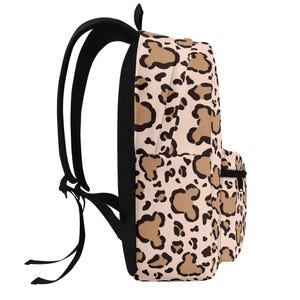 May include: A backpack featuring a light beige base with a repeating animal print pattern composed of tan shapes outlined in black. The bag includes black shoulder straps, a black zipper on the front pocket, and a black handle at the top.