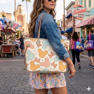 May include: Cream-colored tote bag featuring a pattern of Mickey Mouse heads and bows in pastel colors. The bag has a light brown trim and handles. The background shows a street scene with people and shops.