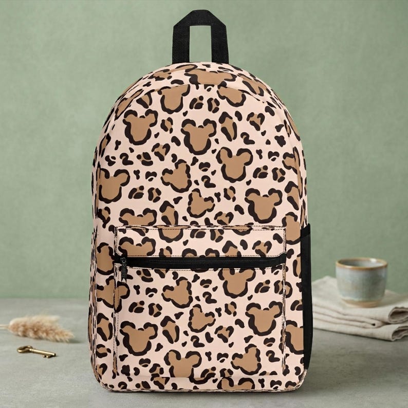 May include: A backpack featuring a light beige background with a repeating leopard print pattern that incorporates brown Mickey Mouse head silhouettes. The bag includes a black top handle, black side panels, and a front zippered pocket with black trim.