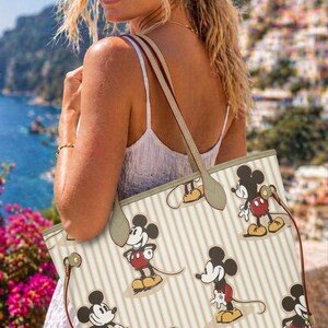 Op de afbeelding: Een beige draagtas met verticale strepen en een herhalend patroon van Mickey Mouse. De tas heeft bruine handvatten en een rode trekkoordsluiting. De achtergrond toont een kustlandschap met gebouwen en water.