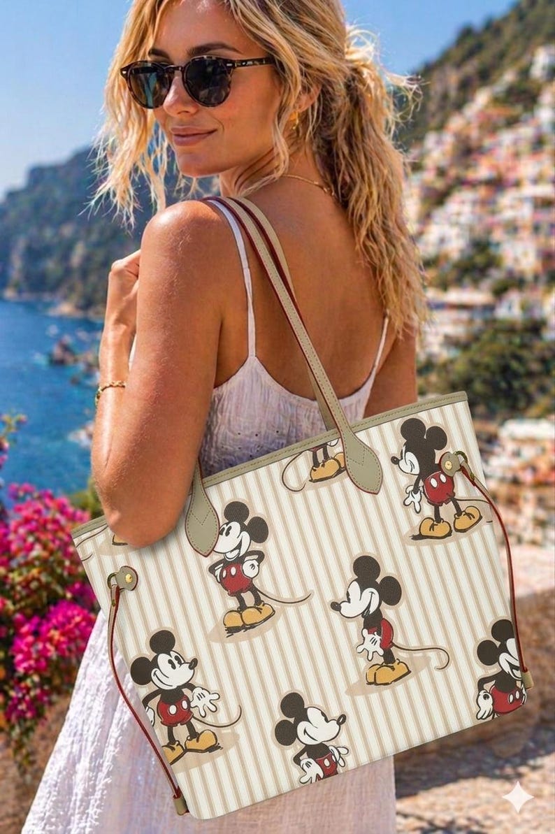 Op de afbeelding: Een beige draagtas met verticale strepen en een herhalend patroon van Mickey Mouse. De tas heeft bruine handvatten en een rode trekkoordsluiting. De achtergrond toont een kustlandschap met gebouwen en water.