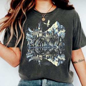 May include: Dark grey t-shirt with a detailed illustration of a castle on a mountain, reflected in a lake. The design includes a snowy mountain range, trees, and a flying owl. A casual wear item.