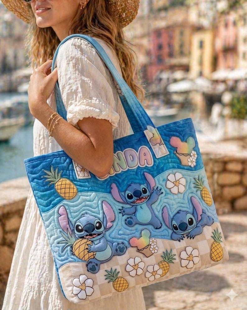 May include: A blue quilted tote bag featuring the character Stitch from Lilo & Stitch. The bag has a beach scene with Stitch, pineapples, and ice cream cones. The word "NDA" is visible on the bag.