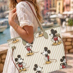 Op de afbeelding: Cr&egrave;mekleurige tote bag met een herhalend patroon van Mickey Mouse in verschillende poses. De tas heeft verticale beige strepen, rode trekkoorden en een beige handvat. De tas wordt gedragen door een persoon die een witte jurk draagt.