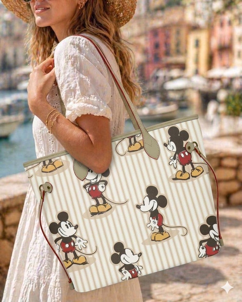 Op de afbeelding: Cr&egrave;mekleurige tote bag met een herhalend patroon van Mickey Mouse in verschillende poses. De tas heeft verticale beige strepen, rode trekkoorden en een beige handvat. De tas wordt gedragen door een persoon die een witte jurk draagt.