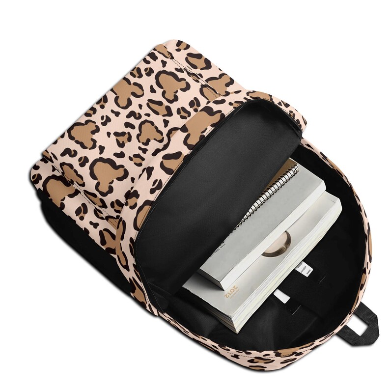 May include: A backpack featuring a beige and brown leopard print pattern on the exterior. The bag is open, revealing a black interior lining and two white notebooks stacked inside. The backpack has a front zippered pocket and black straps.