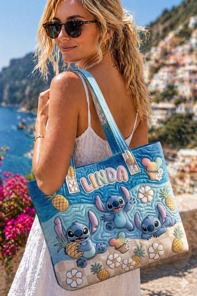 May include: Blue tote bag featuring Stitch characters, pineapples, and flowers. The bag has a quilted design with the name "LINDA" in pastel colors. The bag is carried over the shoulder.