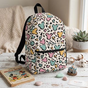 K&ouml;nnte beinhalten: Ein Rucksack mit cremefarbenem Hintergrund und einem wiederkehrenden Leopardenmuster, durchsetzt mit pastellfarbenen rosa, gelben und t&uuml;rkisfarbenen Mickey-Maus-Kopf-Silhouetten. Der Rucksack hat eine Fronttasche mit Rei&szlig;verschluss und schwarze Tr&auml;ger.