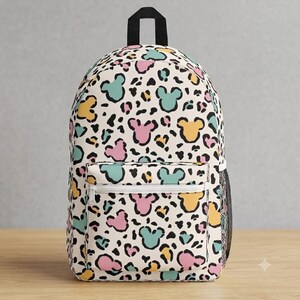 K&ouml;nnte beinhalten: Ein Rucksack mit cremefarbener Basis und einem sich wiederholenden Tiermuster, das mit stilisierten Maus-Kopf-Silhouetten in Pastellrosa, T&uuml;rkis und Gelb durchsetzt ist. Die Tasche verf&uuml;gt &uuml;ber eine Fronttasche mit Rei&szlig;verschluss und einen schwarzen Tragegriff.