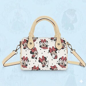 Op de afbeelding: Witte handtas met een patroon van Minnie Mouse-illustraties met rode strikken en zwarte stippen. De tas heeft een cr&egrave;mekleurige handgreep en schouderriem, met goudkleurige hardware.
