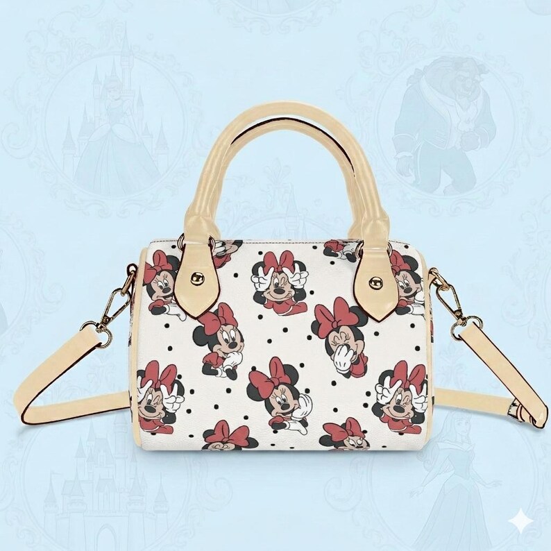 Op de afbeelding: Witte handtas met een patroon van Minnie Mouse-illustraties met rode strikken en zwarte stippen. De tas heeft een cr&egrave;mekleurige handgreep en schouderriem, met goudkleurige hardware.