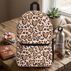 May include: A backpack featuring a light beige background with a brown and black leopard print pattern that incorporates Mickey Mouse head silhouettes. The bag includes a black top handle, a front zippered pocket, and black side accents.