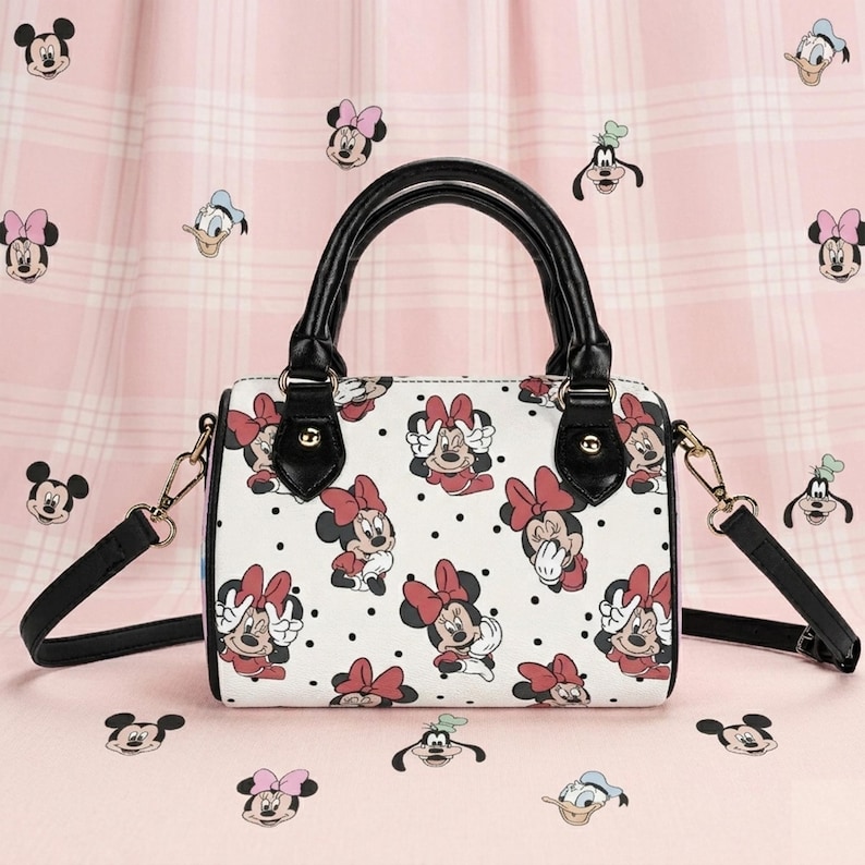 Op de afbeelding: Witte handtas met een patroon van Minnie Mouse met rode strikken, zwarte stippen en zwarte handvatten. De tas heeft een afneembare schouderriem en goudkleurige hardware. Ongeveer 25 cm lang.