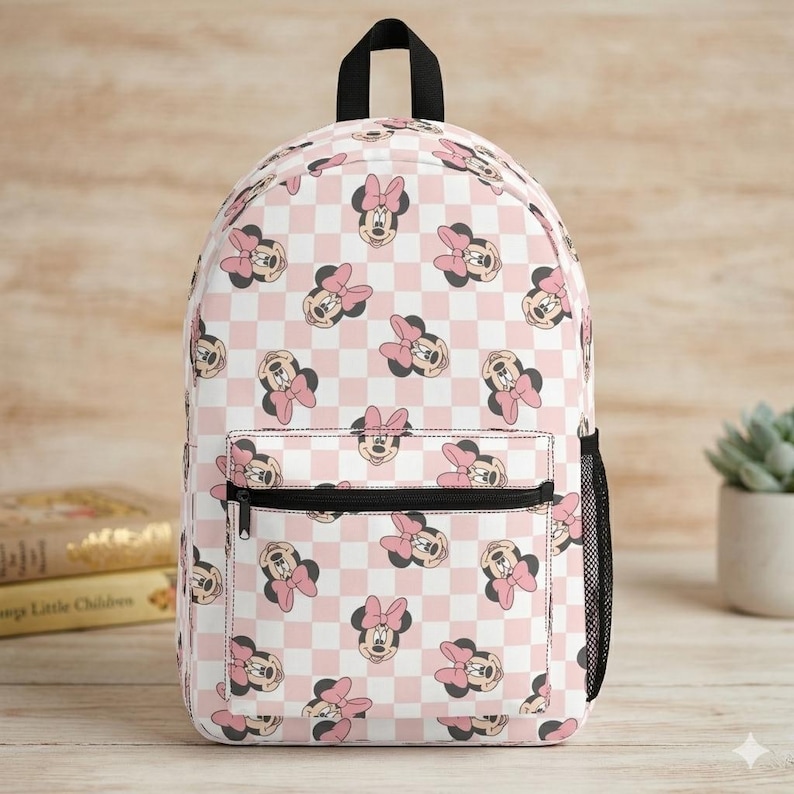 Peut inclure: Un sac &agrave; dos avec un motif r&eacute;p&eacute;titif de visages de Minnie Mouse avec des n&oelig;uds roses sur un fond &agrave; carreaux rose clair et blanc. Le sac comprend une poign&eacute;e sup&eacute;rieure, une poche avant zipp&eacute;e et une poche lat&eacute;rale en filet noir.