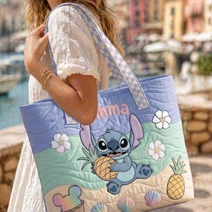 May include: A light blue quilted tote bag with a cartoon character holding a pineapple. The bag has a checkered strap and the name "mma" in pink. Decorative elements include flowers, seashells, and a Mickey Mouse ice cream.