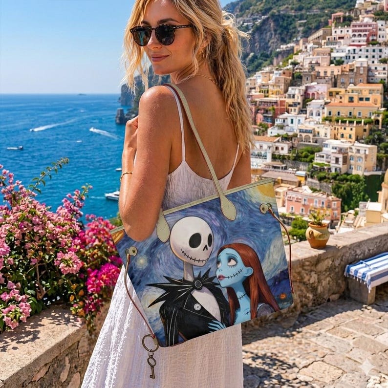 Puede incluir: Un bolso tote con una ilustraci&oacute;n impresa de Jack Skellington y Sally de Pesadilla antes de Navidad sobre un fondo azul de noche estrellada, con asas finas de color beige y un peque&ntilde;o dije de llave de metal colgando.