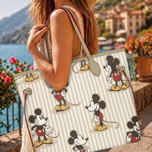 Op de afbeelding: Cr&egrave;mekleurige draagtas met verticale beige strepen en meerdere Mickey Mouse-illustraties. De tas heeft rode en beige bandjes en afwerking. De achtergrond toont een schilderachtig uitzicht op een waterkant met gebouwen en bloemen.