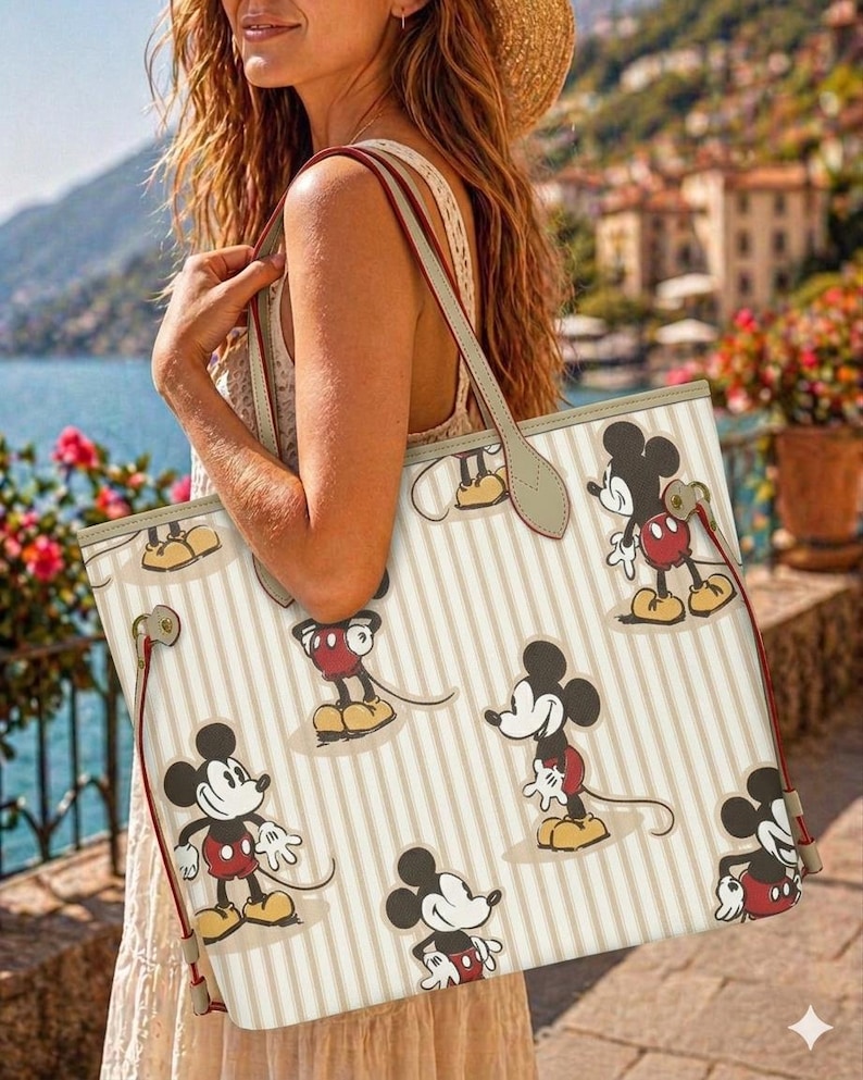 Op de afbeelding: Cr&egrave;mekleurige draagtas met verticale beige strepen en meerdere Mickey Mouse-illustraties. De tas heeft rode en beige bandjes en afwerking. De achtergrond toont een schilderachtig uitzicht op een waterkant met gebouwen en bloemen.