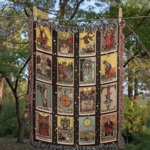 May include: A dark brown woven throw blanket featuring tarot card designs. Each card has a unique illustration and title, including "The Sun," "The Fool," and "The Lovers." The blanket is decorated with fringe and hung with wooden clothespins.