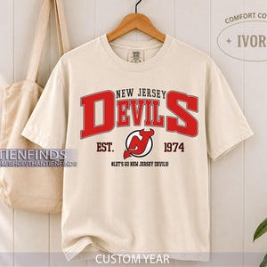 Könnte beinhalten: Ein elfenbeinfarbenes Kurzarm-T-Shirt mit dem roten Schriftzug NEW JERSEY DEVILS, dem Team-Logo sowie dem Text EST. 1974 und #LET'S GO NEW JERSEY DEVILS! Das T-Shirt hängt an einem Holzkleiderbügel neben einer beigen Stofftasche.