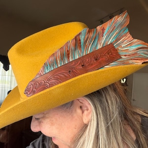 May include: A mustard yellow felt hat featuring a wide brown leather band with intricate floral tooling. The band is accented by a large, fan-shaped decorative element made of layered, fringed leather in shades of brown and turquoise blue.