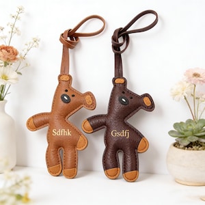Può includere: Due portachiavi in pelle a forma di orsetto. Uno è marrone chiaro, l'altro marrone scuro. Entrambi hanno dettagli beige sulle zampe e sulle orecchie, con occhi neri e bocca cucita. Ognuno ha un anello in pelle per l'attacco e testo sul corpo.