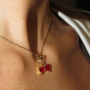 May include: A gold-colored necklace with a cherry pendant. The pendant features two red heart-shaped gems and a small, sparkling bar. The necklace is worn around the neck.