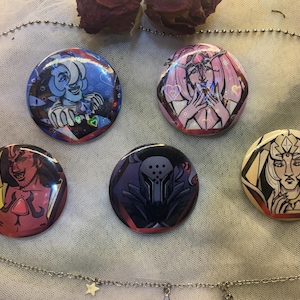May include: Five round pinback buttons with colorful illustrations. The buttons feature various character designs in shades of blue, pink, red, and black. The buttons are arranged on a light-colored fabric with a silver chain and dried flowers.