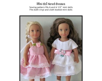 May include: Two dolls wearing tiered dresses. The doll on the left is wearing a pink and white gingham dress with white lace trim. The doll on the right is wearing a white dress with white lace trim and a pink ribbon around the waist. Both dolls are wearing white shoes.