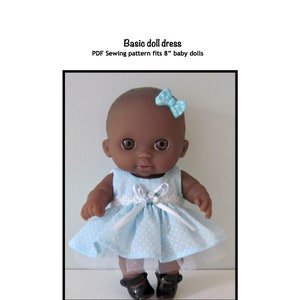 Puede incluir: Un patrón de costura PDF para un vestido de muñeca de 20 cm. El patrón es para un vestido simple, azul claro con lunares blancos y un borde de encaje blanco. La muñeca lleva el vestido y tiene piel morena y zapatos negros.