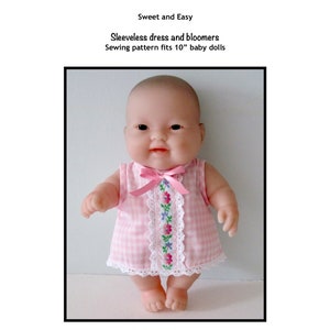 Pode incluir: Um vestido sem mangas de vichy rosa e branco com uma borda floral e um laço rosa. O vestido é projetado para uma boneca de 25 cm. A boneca está vestindo o vestido e tem uma tez clara e cabelos escuros.