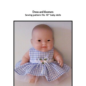 Puede incluir: Un vestido de cuadros vichy azul y blanco con un ribete de encaje blanco y un acento de flor rosa. El vestido está diseñado para una muñeca de bebé de 25 cm. El texto "Dress and bloomers Sewing pattern fits 10" baby dolls" y "Kathy Anne Designs for dolls" es visible en la imagen.