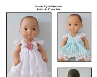May include: Two dolls wearing summer outfits. One doll is wearing a white dress with a pink flower and lace trim. The other doll is wearing a blue and white gingham dress with lace trim. The text "Summer top and bloomers" and "Pattern fits 8" baby dolls" is visible at the top of the image. The text "Kathy Anne Designs for dolls" is visible at the bottom of the image.