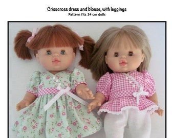 May include: Two dolls wearing handmade outfits. The doll on the left is wearing a green dress with pink flowers and a white ribbon. The doll on the right is wearing a pink and white gingham blouse with white leggings. The caption reads "Crisscross dress and blouse, with leggings. Pattern fits 34 cm dolls. Kathy Anne Designs. For dolls."